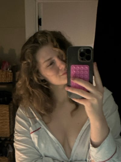 A female individual is depicted in what appears to be an indoor setting, capturing her reflection using a smartphone. The subject has medium-length, wavy, light brown or auburn hair. She is wearing a light-colored, possibly white or light grey, button-up shirt featuring red piping along the collar and cuffs, which is unbuttoned to reveal her upper chest. Her face is partially obscured by the phone, showing a soft expression with eyes seemingly downcast or closed. She holds a dark-cased smartphone in her right hand, which features a prominent multi-lens camera array and a pink, multi-bubble fidget accessory attached to its back. The background to the left reveals a light-colored wall, a dark door hinge, and a section of a woven storage unit containing indistinct items. The background to the right is largely dark and unidentifiable. The location cannot be ascertained from visual evidence within the image.