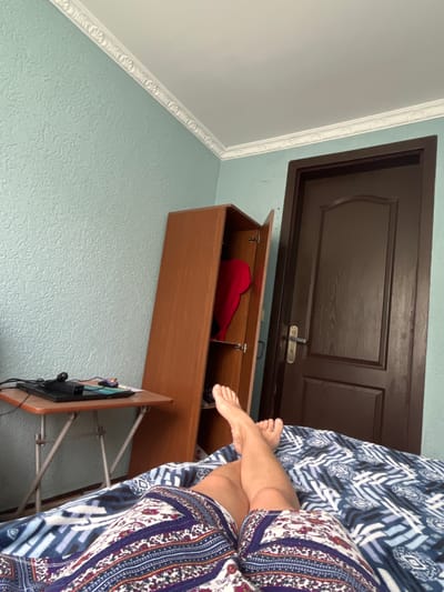 The image depicts an indoor scene, likely a bedroom, located in ღრმაღელის თემი, Georgia. In the foreground, a person's legs and feet are visible, lying on a bed covered with patterned bedding. The person is wearing patterned shorts. To the left, a small, portable table with a laptop and computer mouse is positioned. Behind the table, a tall, open wooden cabinet or wardrobe stands, with a red garment visible inside. To the right, a dark brown wooden door is present. The walls are painted a light blue, with textured wallpaper and white decorative molding along the ceiling. The overall impression is that of a casually occupied personal living space.