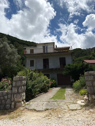 A two-story house with a light-colored facade is nestled into a steep, lush green hillside, under a bright blue sky with scattered white clouds. The house features multiple windows, some fitted with closed dark wooden shutters. An upper-floor balcony with a somewhat rusted metal railing stretches across the facade, and a satellite dish is mounted nearby. Dense foliage, including various trees and shrubs, completely covers the hillside behind and around the house, with an olive tree visible in the lower left.
A concrete pathway leads up to the house from the right, bordered by rough-hewn stone walls. Pink flowering bushes add a touch of color near these walls in the foreground. To the right of the path, a gravel surface is visible. The overall scene suggests a tranquil, natural environment, characteristic of a rural or semi-rural setting like Bratešići, Montenegro. The clear daylight indicates it is daytime. There are no people or visible text in the image. The image is presented with a 90-degree rotation.