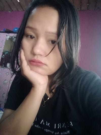 A young woman rests her chin on her hand, gazing forward with a pensive expression. She has dark hair and wears a black t-shirt with white lettering. A delicate gold necklace adorns her neck. In the background, a patterned cabinet with a blue ball on top can be seen. The walls are painted in shades of purple and pink.