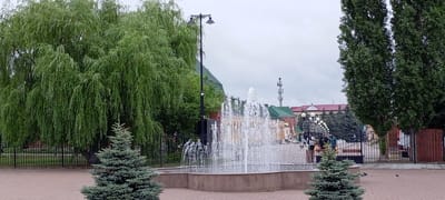 The image shows a public park or square in Kirsanov, Russia, on an overcast day. A large weeping willow tree dominates the left side of the frame, with its long branches reaching towards the ground. In the center, a multi-tiered fountain sprays water upwards. To the right of the fountain, two young evergreen trees are visible. Behind the fountain, several people can be seen, some standing and others walking, near buildings with red and green roofs. A tall, slender street lamp with two lights stands to the left of the fountain, and another similar lamp is partially visible on the far right. The ground is paved with brickwork. In the background, a cellular tower and various trees, including tall, dark green trees on the right, complete the scene.