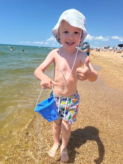 The image shows a young boy at the beach in Blagoveschenskaya, Russia, on a sunny day. He is standing in shallow water, wearing a bucket hat and patterned swim trunks. The boy is holding a blue bucket and has his thumb up, giving a thumbs-up gesture. Sand is visible on his skin and hands. The background shows the sea, a sandy beach populated with people, and a clear blue sky with some clouds. The water is transparent, with small shells and pebbles visible on the seabed.