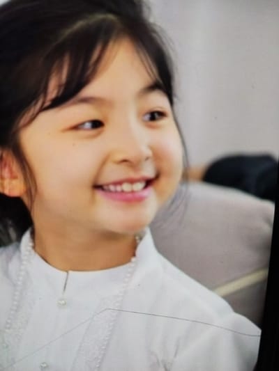 This is a close-up media file of a young East Asian girl. She has dark hair, pulled back from her face, and is looking to her right with a wide, joyful smile that reveals her teeth. Her skin is fair, and her cheeks are rosy. She is wearing a white collared shirt adorned with a pearl necklace and pearl buttons. The background is blurred, showing a light-colored surface that could be upholstery or a wall, and a darker, out-of-focus shape behind her to the right. The lighting appears to be soft and even, suggesting an indoor setting. The overall impression is one of happiness and innocence. There is no visible text in the image.
