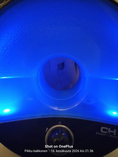 This is a close-up, interior shot of what appears to be a humidifier or air purifier. The device is predominantly illuminated by a bright blue light, creating a vibrant and somewhat futuristic aesthetic. The top surface of the device shows a textured pattern, possibly from condensation or the material itself, and is a deep blue color. In the center, there's a circular opening, a darker void within the blue, through which a dimly lit interior space can be vaguely discerned. The lower portion of the device is dark and features a control dial with markings and a power symbol. A logo reading "CH Cooper & Hunter" is visible on the right side of the dark section. At the bottom of the image, overlaying text in white indicates "Shot on OnePlus," "Pikku kakkonen," and a date and time: "16. kesäkuuta 2026 klo 21.36." The date is in the future, suggesting this may be a timestamp from a test or a manipulated image. There are no people or discernible external environmental elements like cityscapes or landmarks visible, making it impossible to determine the specific location in Tampere, Finland, or any other city.