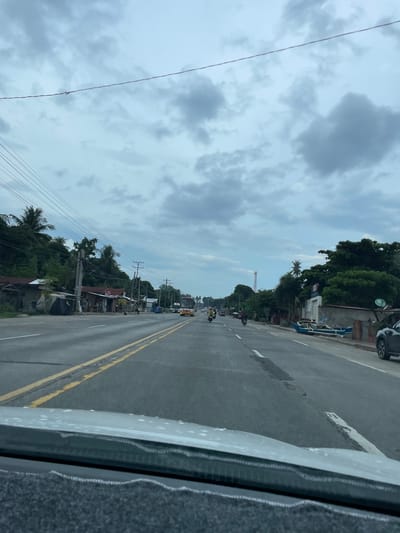 The image shows a view from inside a vehicle, likely a car, driving along a two-lane road in Santa Cruz, Philippines. A yellow double-line divides the lanes. There are trees, houses, and utility poles lining the side of the road. Other vehicles, including motorcycles and a bus, are visible in the distance. The sky is overcast with grey clouds. A tall crane or communication tower is visible in the distance.