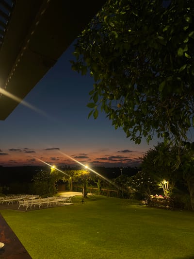 The image shows an outdoor event space at dusk in Beit Hanan, Israel. Rows of white chairs are set up on a grassy lawn, facing a wooden platform adorned with string lights. A tree with green leaves and fruit is in the foreground on the right, and another tree with white flowers is visible behind the chairs. The sky is a gradient of deep blue to orange, with scattered clouds. Lights are illuminating the scene. There are no people visible.