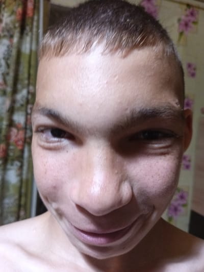 A close-up, slightly blurry, front-facing photograph shows the face of a young male with short, dark hair. The subject is looking directly at the camera with a slight smile. The background consists of a patterned curtain with floral motifs on the left and a wall with a similar pattern on the right. The lighting appears to be artificial and from the front. The image was taken indoors. There is no discernible indication of the location being in Tbilisskaya, Russia.