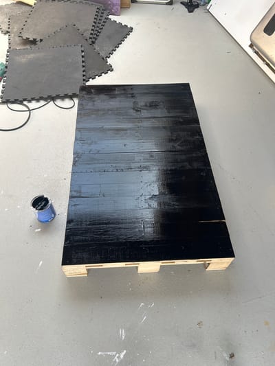 A black, glossy wooden surface, likely a tabletop or a similar flat object, is positioned on a concrete floor. The surface is constructed from several wooden planks laid side-by-side, with visible grain and some imperfections. The edges of the wooden object reveal a lighter wood material, suggesting it's a composite structure or has a wooden frame.
To the left of the wooden surface, a pile of interlocking foam mats, typically used for exercise or play areas, are stacked. These mats are dark gray with a textured surface and interlocking edges. A black cord, possibly an electrical cable, is visible on the floor, partially coiled and extending towards the bottom of the frame.
In the lower right corner of the image, an open can of black paint rests on the concrete floor. The can has a blue label with some white text, but it's largely obscured. Splatters of white paint are visible on the concrete floor in various areas, indicating a workspace or a recent painting project.
The overall setting appears to be an indoor space with a concrete floor, possibly a garage or workshop. The lighting suggests it could be daytime, but there are no direct views of windows or outdoor elements to confirm. The scene conveys a sense of ongoing DIY or crafting activity.