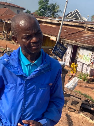 This image captures an outdoor street scene in Amaseri, Nigeria, under bright daylight. In the foreground, an adult Black male with a bald head and stubble-like facial hair faces the viewer, displaying a slight smile. He is visibly sweaty on his forehead and face. He wears a bright blue polo shirt beneath a royal blue zip-up jacket, which has a "SPRAYWAY" logo on the right chest. His right hand is partially visible in the lower right.
The background reveals a bustling environment with multiple structures featuring aged, rust-colored corrugated metal roofs. A light-colored utility pole stands vertically in the mid-ground, partially obscuring the buildings. To the right, a black sign with white text advertises "LAND SURVEY / NEW NUMBER / AND / REGISTRATION" with a phone number (08033549259). Adjacent to this, a large white banner with green text displays "bet9ja" and a soccer ball logo, indicating a sports betting shop. A child wearing a yellow t-shirt and a light-colored skirt walks from left to right in front of the "bet9ja" banner, and another individual is partially visible further to the right.
In the distant background, a distinctive building with an angular, multi-peaked roof is visible, along with a satellite dish on a nearby roof. Palm trees and other tropical vegetation punctuate the sky. The ground is unpaved, appearing as reddish-brown earth with patches of green grass. A white bag, a small wooden stool, and dark metal pots or basins are scattered on the ground in the mid-foreground.