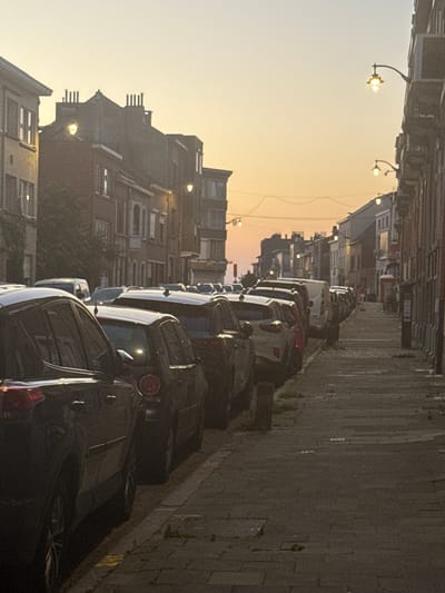 The image depicts a street scene in Molenbeek-Saint-Jean - Sint-Jans-Molenbeek, Belgium, during what appears to be sunrise or sunset, indicated by the warm, hazy sky. The street is lined with parked cars on the left side and buildings on both sides. The architecture of the buildings suggests an urban European setting, with multi-story structures featuring pitched roofs and chimneys. Streetlights are visible along the street, some illuminated. On the right side of the street, there is a sidewalk with a cobblestone pattern. The cars are parked in a line, and their reflections are visible on the pavement. The foreground shows the rear of a dark-colored SUV. The overall atmosphere is one of a quiet urban morning or evening. There are no visible people in the image.