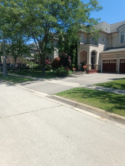The image depicts a suburban street in Oakville, Canada, on a clear, sunny day. In the foreground, a section of a paved road and a concrete sidewalk are visible, with a grass verge and a curb separating them from the property line. The street features a painted line dividing lanes.

The main subject of the image is a large, upscale house with a light-colored stucco exterior. The house has a prominent second-story balcony with an ornate railing, supported by columns that frame an arched entryway. The house is flanked by two large, dark red garage doors. A number "85" is visible above the right garage door.

Lush green trees, some with mature foliage, are present in front of the house, casting dappled shadows on the lawn. Red ornamental trees and bushes add color to the landscaping. A street light pole with a "no parking" sign is visible on the left side of the image. Adjacent properties and houses are partially visible behind the trees and in the background. The environment is characteristic of a well-maintained residential neighborhood in Oakville. There are no people or animals visible in the scene. The scene appears to be static and undisturbed.