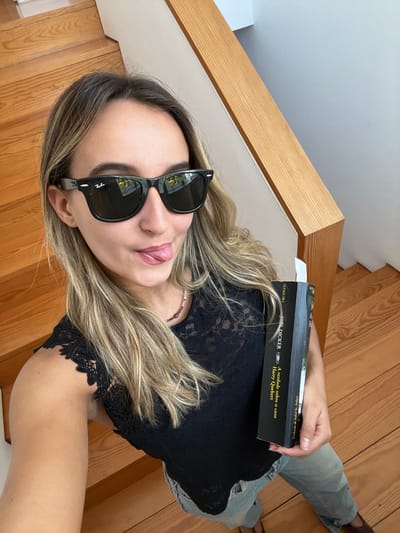 The image captures a female subject positioned on an indoor wooden staircase in what is stated to be Oliveira de Frades, Portugal. The subject, an individual with light brown, wavy hair, is wearing black Ray-Ban Wayfarer-style sunglasses, a black lace sleeveless top, and light-wash blue jeans. A subtle, multi-colored beaded necklace is visible. She is taking a selfie, holding the camera from a slightly elevated perspective, and looking directly towards the lens with her tongue partially extended from her mouth. In her left hand, she holds a black-spined book titled "A verdade sobre o caso Harry Quebert" by Joël Dicker, with "ALEGGUARA" visible as the publisher. The background consists of natural-grain wooden stair treads and risers, a light-colored wooden handrail or newel post, and plain light-toned walls.