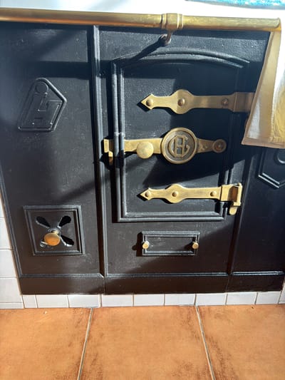 The image displays the front of a dark, likely cast-iron, wood-burning stove or oven, located in Parres, Spain. The surface is textured and appears to be painted or enameled black. A prominent horizontal brass-colored bar serves as a rail across the top. Below this, the front panel features multiple brass-colored hardware elements.

Three decorative brass-colored latches are visible on the right side of the main door panel. The central latch is circular with an embossed stylized "G" or "C G" monogram. Below these, a small rectangular plate with two circular brass-colored knobs is present. On the lower-left panel, a square-framed vent with a quatrefoil-shaped opening houses a central brass-colored knob, likely a damper control. An embossed emblem resembling a stylized "LH" within a house or shield shape is located on the upper-left panel.

A light-colored fabric, possibly a towel, is draped over the right end of the top brass rail. The stove rests on a base of white rectangular tiles, which transition to a terracotta-colored tiled floor. Direct lighting from the upper left casts distinct shadows across the textured surface of the stove. No individuals or active interactions are present in the scene.