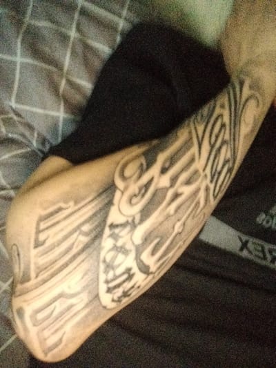 An arm with a large, stylized tattoo is lying on a surface with a gray and white checkered pattern. The tattoo is black and gray, featuring intricate line work and lettering. A dark-colored shirt is partially visible behind the arm. No indication of being in Gus-Khrustalny, Russia.