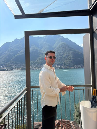 A man stands on a balcony in Pognana Lario, Italy, looking out at a sun-drenched lake and mountains. He is wearing a light-colored, unbuttoned shirt and dark pants. The balcony has a metal railing and a white trash can containing two closed umbrellas. The lake sparkles under the bright sun, and a town is visible on the opposite shore, nestled at the foot of lush green mountains. The sky is clear and blue.