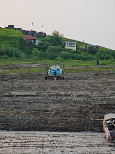 A wide shot captures a rural scene in Potapovo, Russia, on a clear day, likely in the late afternoon or early evening given the soft, diffused light. The foreground is dominated by a dry, gravelly riverbed, with the calm, rippling water of a river visible at the bottom of the frame. A reddish boat, partially submerged, rests at the edge of the river on the right side of the image.
Further back, a small, light blue motorboat is beached on the gravelly shore, resting on a trailer. It is positioned centrally in the frame, facing the viewer. A darker, distant figure stands on the grassy embankment behind the boat, engaged in an unseen activity.
The middle ground features a lush green, sloping hillside dotted with small yellow flowers and scattered trees. Along the crest of the hill, a cluster of simple wooden houses and structures are visible, suggesting a small settlement. Power lines stretch between poles, connecting the dwellings. The architecture of the houses, with their pitched roofs and wooden construction, indicates a rural or remote setting. The overall atmosphere is one of quiet, everyday life in a northern landscape. No visible text is present in the image.