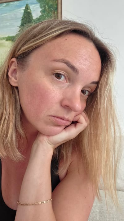 The image shows a close-up, sideways portrait of a young woman with blonde hair. She is lying down, with her head resting on her arm and hand. Her eyes are open and looking towards the camera. She has light-colored skin and her lips are slightly parted. There is a subtle blush on her cheeks. A thin, gold chain bracelet is visible on her arm. The background is blurred, but appears to be an indoor setting with a painting on the wall that depicts a landscape with trees. The lighting suggests it could be daytime. The woman's pose and expression could be interpreted as contemplative or relaxed. The location is identified as Guía de Isora, Spain.