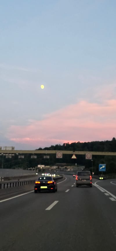 A vertical, low-angle, point-of-view shot captures a highway scene at dusk in Urdorf, Switzerland. The sky is a gradient of pale blue to soft pink with a luminous yellow moon visible in the upper left quadrant. A line of dark green trees and a yellow overhead gantry with traffic signs are visible in the background. The highway has multiple lanes, with white dashed lines marking the boundaries. Two cars are driving away from the camera. The car in the foreground, a black sedan, has its brake lights illuminated. Further ahead, a dark-colored SUV is also traveling away from the viewer. The overall lighting suggests twilight conditions.