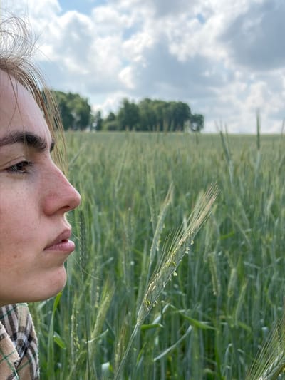 An outdoor, medium close-up shot shows a young person's profile looking to the left from the chest up, in a field of green wheat under a partly cloudy sky. The person's right eye, nose, and mouth are visible, with fine facial hairs and freckles. Their hair, a light brown, is windswept and falls across their forehead and cheek. They are wearing a plaid shirt. The background is a vast expanse of ripening wheat, with a line of trees in the far distance. The image was taken in Gushchino, Russia.