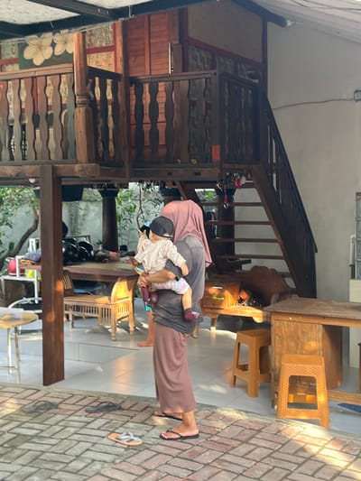 A person, likely a woman wearing a pink headscarf and a long, brown skirt, is holding a young child. The child is wearing a light-colored hooded jacket and a dark baseball cap. They are standing on a brick patio in front of a multi-level wooden structure. The structure features a railing with ornate wooden carvings and a wooden staircase. In the background, there is a table with several black helmets and some greenery. There are also several wooden chairs and stools, as well as a white shelf unit. The scene appears to be outdoors, possibly in a courtyard or a backyard, in Pemenang Timur, Indonesia. The lighting suggests it is daytime, with sunlight casting shadows on the patio.