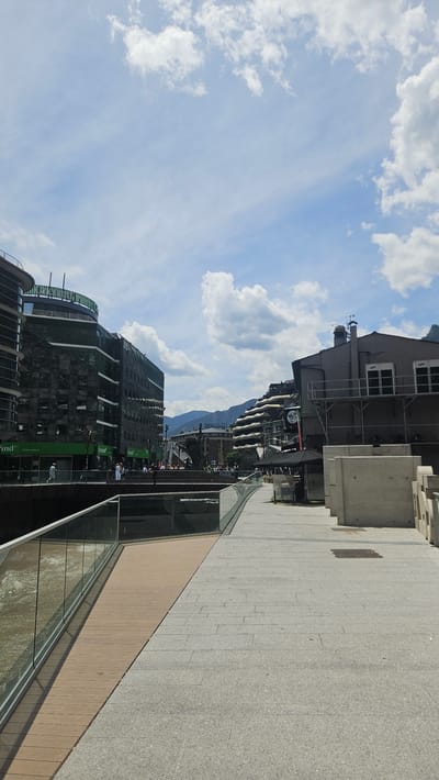 This image depicts a vibrant urban scene in Andorra la Vella, Andorra, specifically along the Gran Valira river. The foreground features a modern pedestrian walkway. On the left, a light brown wooden deck abuts a glass safety barrier, beneath which a brownish-grey river with visible current flows. This wooden section transitions to a wider, light grey paved path composed of rectangular stone blocks with a speckled aggregate finish, extending towards the viewer and into the distance on the right. On the far right, a series of light grey concrete block structures are arranged.
In the midground, to the left, stands a multi-story, modern building characterized by dark, reflective glass or stone facades. Its upper section features a distinctive curved, green-topped structure. Visible signage on its lower level includes "Find" and "Crèdit Andorrà" on green fascia elements. In front of this building, on a lower-level walkway, a few individuals are visible. To the right of the scene, a darker grey building with a pitched roof, a chimney, and two white rectangular external units (possibly HVAC) mounted on its side is present. A metal framework or scaffolding is attached to its exterior. Below this building, a black canopy or large umbrella is situated.
Further into the midground, centrally, an elevated public space or plaza is populated by numerous individuals. A large, dark-colored statue or sculpture is visible within this gathering. In the background, rolling mountains covered with dark green vegetation rise, partially obscured by the urban structures. Several buildings with distinct terraced or step-like architectural designs are integrated into the hillside on the right side of the background. The sky above is a bright, clear blue, adorned with scattered white cumulus clouds, indicating a sunny day.