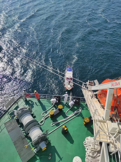 This image captures an overhead view of a maritime pilot transfer operation from a large ship. The scene is dominated by the ship's green deck and the vast expanse of blue, rippling ocean.

On the vessel's deck, several individuals are actively involved in or observing the operation. Three people are dressed in white coveralls, with one facing the pilot boat and another positioned further back. Two other crew members, one in an orange suit with a yellow hard hat and another in a red suit with a white hard hat, are situated near the ship's edge, possibly managing lines or overseeing the transfer. Deck equipment includes a large spool of thick white rope, black and yellow striped bollards, and a metal grating. An orange enclosed lifeboat is visible, suspended from its davit system, which bears the text "SWL 59KN" (Safe Working Load 59 Kilonewtons). Another black object on the deck is marked "SWL 7.2t."

Alongside the ship, a smaller white pilot boat is visible, clearly identified by the red "PILOT" text on its side. It is connected to the main vessel by several white ropes, which appear to be guiding lines or fenders. At least two people are aboard the pilot boat, one in red seemingly at the controls.

The ocean exhibits waves and sparkling reflections from strong sunlight, indicating clear weather conditions during the day. The bright light also casts distinct shadows on the ship's deck. This functional scene depicts a common procedure in maritime navigation, occurring in the waters near Kunak, Malaysia.