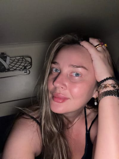 A fair-skinned person with blondish-brown hair, wearing jewelry including rings and bracelets, is positioned close to the camera, looking at the viewer. The background includes a wall, a metal folding chair or drying rack, and a hint of dark clothing. The image's orientation is tilted. The location, Krasnyi Yar, Russia, cannot be confirmed from the image content.