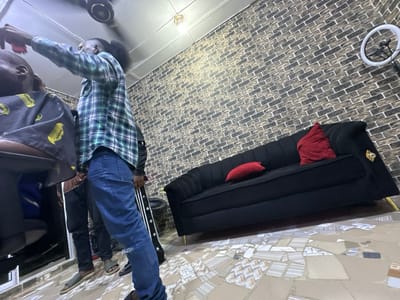 Inside a barbershop, a barber is giving a haircut to a customer. The barber wears a green plaid shirt and blue jeans with a distressed knee. Two people wait, wearing casual clothes. The shop features a black sofa with red throw pillows against a brick-patterned wall. A ring light is mounted on the wall. The floor is decorated with a mosaic tile design. A ceiling fan is visible.