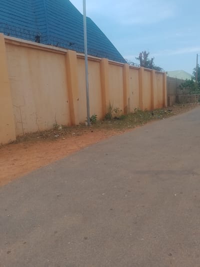 The image shows a residential street in Gindi Nassarawa Kyarmi Galbi, Nigeria. A tall, orange-colored wall with vertical pillars dominates the left side of the frame. Barbed wire is installed along the top of the wall, and a blue-tiled roof is visible above it on the left. The wall extends to the right, where it transitions into a lower, brick wall. Sparse vegetation, including weeds and some small plants, grows along the base of the wall. Debris, such as plastic bottles and paper, is scattered in this area. A paved road runs along the right side of the image, separating the viewer from the wall. A light pole stands near the wall on the right. The sky is overcast.