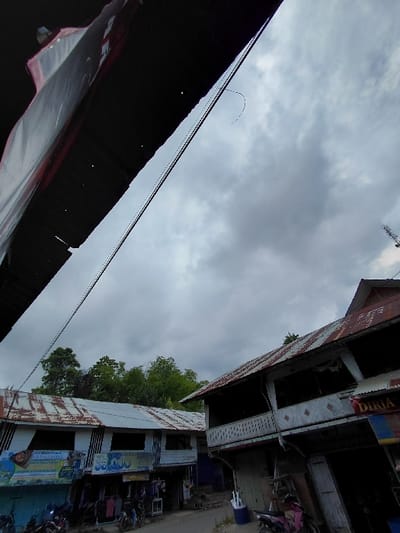 A street scene under a cloudy sky features buildings with aged corrugated metal roofs. A dark awning occupies the lower-left. The buildings line both sides of a narrow street, populated with parked motorcycles and possibly people. Green trees are visible behind one of the structures. Power lines stretch across the sky between the buildings, adding to the urban feel. The camera is angled upwards, emphasizing the height of the structures and the expansive sky.