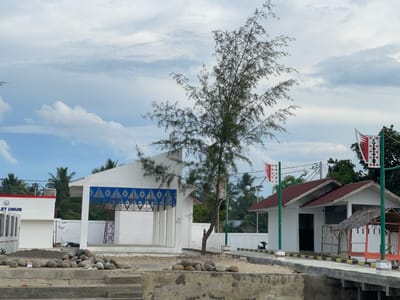 The image depicts an outdoor scene in Lancok, Indonesia, likely during the daytime given the bright overcast sky. The weather appears cloudy. The setting is a coastal or waterfront area with buildings and a pathway.
In the foreground, there is a concrete structure with steps leading up to a sandy area. A pile of smooth, grey stones is scattered on the sandy ground. Beyond this, a white building with an open-air section and decorative blue and gold patterned panels is visible. To the right, another small white building with a red-tiled roof stands. A small, thatched-roof hut is attached to this building. A concrete pathway with yellow and black striped borders runs alongside the buildings. A motorcycle is parked near the pathway.
A tall, slender tree with sparse, needle-like leaves stands in the middle ground. Two flagpoles with distinctive red and white flags are positioned in front of the buildings. Palm trees and other vegetation are visible in the background, suggesting a tropical location.
There is visible text on the white building to the left, reading "LET UMUM". The overall impression is of a developing or recently constructed area, possibly a public facility or tourist spot. The scene is calm and devoid of people.