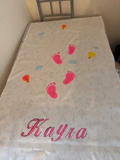 The image displays a white fabric item, likely a blanket or sheet, adorned with vibrant, personalized designs. The base fabric features a subtle, light grey floral and leaf pattern. The most prominent elements are five pink footprints, each containing a word or number written in white. These include "Four", "Love", "Three", "One", and "Two", read from left to right if the fabric were oriented upright.
Interspersed with the footprints are abstract, softly colored shapes: light blue ovals and yellow/orange designs resembling hot air balloons or lanterns. On the right side of the fabric, a large name, "Fayega" (or possibly "Faye-ga"), is written vertically in elegant pink cursive script with a black outline, suggesting a personalized item for a child.
The fabric is laid flat, possibly on a bed or cot, as indicated by a grey metal bed frame or railing visible along the left side of the image. A plain, light-colored wall forms the background on the upper left. The overall aesthetic is gentle and child-centric, implying the item is a keepsake, potentially celebrating a birth or early milestones. The absence of people indicates a focus on the object itself, presented in an indoor setting, likely a nursery or bedroom in Bearpark, United Kingdom.