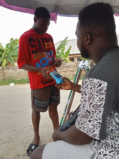 Two men stand on a dirt road in Sapele, Nigeria, located at the coordinates 5.86 latitude and 5.701 longitude. The man on the left is wearing a red t-shirt with a blue and white graphic design, black and blue shorts, and black flip-flops. He is holding a black smartphone in his left hand and a blue and black POS machine in his right hand. The man on the right is wearing a black and white patterned shirt and gray shorts. His left hand is holding the POS machine, his right hand is resting on his leg. There is a white plastic bag on the ground between the two men. A leafy green plant grows in front of a concrete wall, and a red awning is above them. The image was taken in broad daylight.