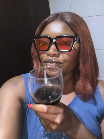A woman with red-tinted sunglasses and reddish-brown hair holds a glass with liquid inside. She wears a blue top. Her nails are painted red. The background consists of what appears to be a tiled wall on the top-left and a dark-colored flat surface towards the bottom. The image is oriented sideways.