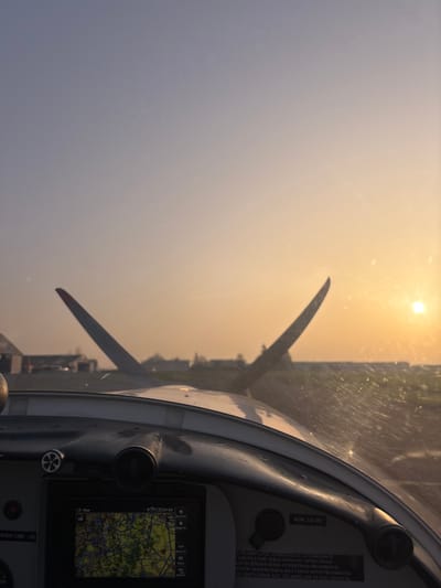 The image presents a first-person view from within the cockpit of a light aircraft at Toussus-le-Noble Airport, France, under the warm, hazy light of either sunrise or sunset. The sun, a bright orange disc, is visible low on the right horizon, casting a distinct glow across the sky, which transitions from pale blue overhead to orange-yellow at the horizon.

The aircraft's grey and white dashboard occupies the lower portion of the frame. On the left, a digital display shows a navigation map with road networks and terrain, indicating the time "17:53." To the right of the display, an "ALPA 12V DC" label is visible, along with a partial advisory stating "THIS AIRPLANE IS APPROVED IN ACCORDANCE WITH..." and referencing "DAY/NIGHT OPERATIONS" and "LIMITED AEROBATICS" with other instructions pertaining to the aircraft flight manual. Vents and control knobs are also present on the dashboard.

Through the windshield, which shows minor smudges and reflections, parts of the aircraft's white cowling and two upward-curved propeller blades are visible, silhouetted against the sky. Beyond the aircraft, a paved surface, consistent with an airport taxiway or runway, extends into the distance. On the far left, in the middle ground, several hangars or airport buildings are discernible. A distant line of vegetation or trees is visible on the far right. No individuals are present in the scene.