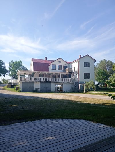 The image depicts a multi-story white building with a red metal roof, situated in Tingstäde, Sweden. The building features a prominent wrap-around balcony on the first floor, supported by white pillars. Several wooden doors are visible on the ground level. A spiral staircase is attached to the right side of the building, leading up to the balcony. In front of the building, there is a paved pathway and a grassy area. A white umbrella and a table with chairs are set up on the balcony. Trees are present on either side of the building, and the sky is blue with some wispy clouds. The foreground is dominated by a wooden deck.
