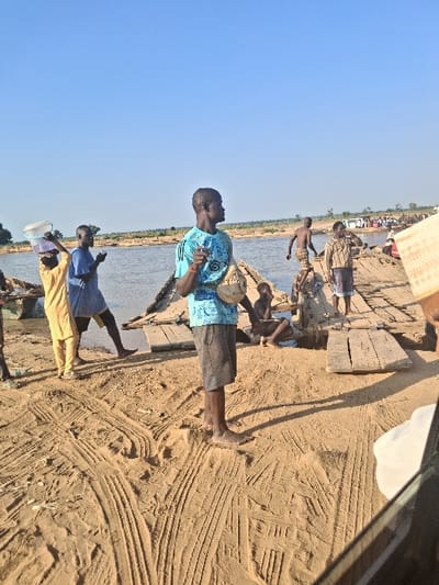 The image depicts an outdoor scene at a river crossing in Nemnei, Nigeria, under a clear blue sky. The foreground consists of light brown sand exhibiting numerous tire tracks. In the center, a dark-skinned male stands barefoot, wearing a light blue patterned short-sleeved shirt and dark shorts, holding a small light-colored pouch and gesturing with his right hand. To his left, a person in a yellow garment carries a white and a purple plastic bucket on their head, facing towards the water. Behind them, another dark-skinned male in a blue shirt stands near the water's edge.
A makeshift wooden bridge or dock, constructed from rough planks, extends from the sandy bank into a wide body of water occupying the mid-ground. Several individuals are present on and around this structure. A shirtless dark-skinned male is stepping onto the wooden structure from the water, while another dark-skinned male in a plaid shirt and wrapper stands further along the planks, holding an object to his mouth. A child or small individual sits on the sand adjacent to the wooden structure.
The distant riverbank features sparse vegetation and more indistinct figures, suggesting further activity. A portion of what appears to be a vehicle's frame and a woven object are visible in the bottom right corner. The scene is brightly lit by natural sunlight, casting shadows to the left.