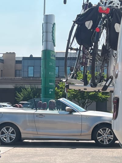 A silver convertible BMW is parked on a paved surface, with its top down. Two individuals are seated in the car, one in the driver's seat and another in the passenger seat. A white vehicle with a bicycle rack is partially visible to the right of the BMW. The bicycle rack holds multiple bicycles, some with panniers. In the background, a tall, green cylindrical structure with white trim stands prominently. This structure appears to be a gas station sign, displaying text such as "ULTRA," "24/24," "car," "euro," and "ULTRA 98." Behind the gas station sign, a building with dark-tinted windows and a concrete facade is visible. Lush green trees and foliage surround the area, suggesting an outdoor environment. The scene is bathed in sunlight, indicating a clear day. This image was captured in Weiswampach, Luxembourg.