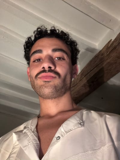 This image captures a close-up, low-angle portrait of an adult male within an indoor setting in Mauléon-d'Armagnac, France. The subject has dark, curly hair, brown eyes, an olive complexion, and a dark, trimmed mustache and beard. His lips are slightly pursed, and he is looking directly forward. He is wearing a light-colored, possibly white or off-white, collared garment featuring visible stitching and metallic snap fasteners, partially unfastened to reveal his upper chest and neck. The background consists of a white or light-colored ceiling or wall with horizontal paneling or joists visible in the upper left. A prominent, rough-hewn, dark wooden beam is positioned diagonally across the upper right portion of the frame. The scene is illuminated by diffused overhead lighting.