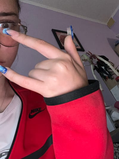 The image shows a close-up of a person's hand and arm. The person is wearing a red Nike jacket with black trim. The hand is positioned with the index and middle fingers extended downwards, forming a V-shape. The fingers have long, artificial nails with a blue and white design. Part of the person's face is visible, including an eye and glasses. The background is a wall painted a light purple color, with a framed picture hanging on it. There are also shelves in the background with some items on them, including what appears to be decorative items and some white packaging. The lighting is dim, suggesting it might be evening or indoors. The overall impression is of a casual, personal photo, possibly taken at home. The visible text is "NIKE". The location context is Chavdar, Bulgaria.