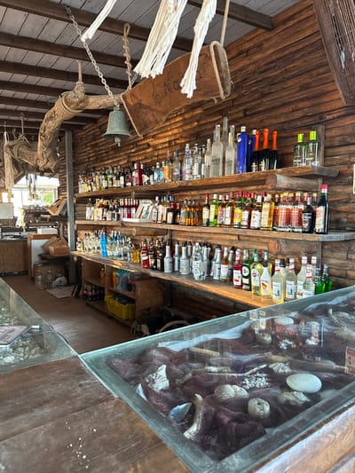 The image shows a bar area with a rustic, wooden interior. The walls and shelves are made of logs, and there are various bottles of alcohol displayed on the shelves. In the foreground, a glass-topped counter displays maritime-themed decorations, including fishing nets, shells, and driftwood. A bell hangs from a chain near the ceiling, along with some white decorative ropes. The overall atmosphere suggests a casual, possibly beachside establishment. The location is identified as a bar in Angelochori, Greece. No people are visible in the image.