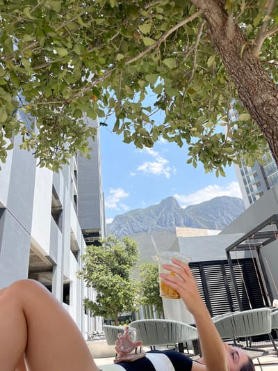 The image shows a woman lying on a lounge chair in a courtyard in the city of Ciudad Santa Catarina, Mexico (latitude: 25.657, longitude: -100.449). She is holding a glass of iced coffee in her right hand and looking up at the sky. The lounge chair has black and white striped cushions. The courtyard has a modern design, with concrete walls and black metal railings. There is a large tree in the foreground, with its branches extending out to the left and right. The tree shades the woman and the lounge chair from the sun. In the background, there is a mountain range and modern apartment buildings. The scene is peaceful and relaxing, with a clear blue sky and lush green vegetation. The woman appears to be enjoying a moment of peace and quiet.