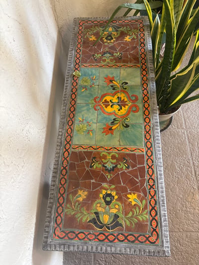 The image shows a mosaic-tiled table with a decorative border. The tabletop is divided into sections, with the central section featuring a teal background with a yellow and orange floral motif. The top and bottom sections of the tabletop have a brown background with green and yellow floral designs. The borders of the table are adorned with a repeating pattern of orange, black, and green geometric shapes. A snake plant in a pot is visible to the right of the table, and a textured wall is on the left. The floor appears to be tiled. The image was taken in Delray Beach, United States of America.