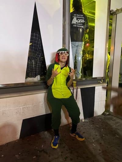 A person in a green and yellow dinosaur costume with shark tooth glasses lies on a black and white tiled surface next to a window reflecting a crowded interior. The window also shows a man wearing a black t-shirt with white writing "NO MATTER THE QUESTION, IBIZA IS ALWAYS THE ANSWER." The person in the dinosaur costume wears blue and yellow sneakers. The location is presumed to be Grau I Platja, Spain.