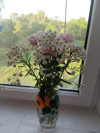 A bouquet of pink and white flowers sits in a clear glass vase on a windowsill. The vase is decorated with a painted design of sunflowers and green leaves. Through the window, a lush green landscape with trees can be seen, suggesting a natural, outdoor setting. The light filtering through the window indicates it is daytime, likely a clear or partly cloudy day. The windowsill is covered with a white textured material. The image has a peaceful and domestic feel, with the flowers bringing a touch of nature indoors. There is no visible text in the image.