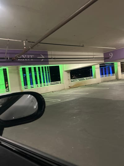 The image depicts a view from inside a multi-story parking garage at night, likely on the ninth level, identified by a prominent white numeral "9" on a purple panel. The location is the parking garage of the Atlantis Casino Resort Spa in Reno, Nevada, United States of America.

The perspective is from within a vehicle, with a portion of a black car side mirror visible in the lower left, reflecting the interior concrete ceiling. The ceiling is off-white, with various exposed brown, white, and black pipes running horizontally. A purple band on a structural beam features partially visible white text "EXIT" with an arrow pointing right.

Through the large, rectangular openings along the exterior wall of the garage, a prominent building facade is visible against a dark night sky. This building exhibits distinct vertical green light strips and horizontal blue light strips, characteristic of the Atlantis Casino Resort Spa. The garage openings are framed by concrete pillars and sills, with a white, multi-bar railing running along the edge of the openings. The concrete floor of the parking garage is visible, dark gray with faint white painted lines indicating parking stalls and concrete wheel stops. No vehicles are present in the visible parking spaces. A blurred reddish object or person is faintly discernible through one of the green-lit openings on the far left.