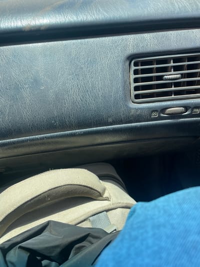 The image shows the dashboard of a vehicle. The dashboard is textured and has an air vent with adjustable louvers and a dial. Next to the dashboard is a backpack with a tan strap and some black material. A blue object is visible above the backpack, possibly clothing. The location is unknown and there is no information about Unguwar Gaya, Nigeria, or any people or interactions.