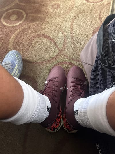 A first-person, overhead shot shows a person wearing a pair of maroon Nike soccer cleats on their right foot and a blue/grey Adidas shoe on their left foot. Both feet are clad in white athletic socks. The person is standing on a carpet patterned with tan and brown circular designs. Part of a grey and black mesh bag is visible next to the right foot. The location is not discernible in the image, and therefore its location in Charentsavan, Armenia cannot be determined.