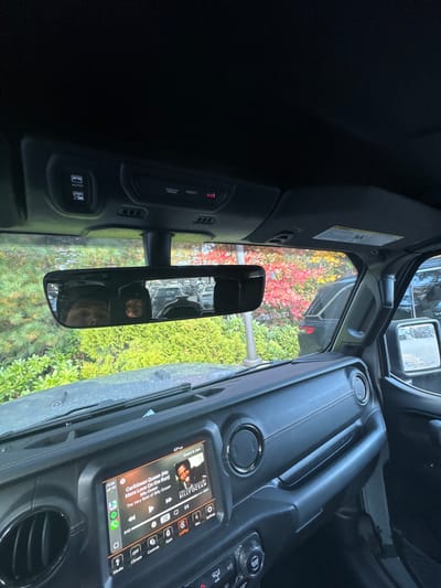 The image depicts the interior of a dark-colored vehicle, possibly a Jeep, looking forward through the windshield. The infotainment screen on the dashboard displays "Caribbean Queen (No More Love On the Run)" by Billy Ocean playing on Spotify, along with a temperature reading of 67°F. The overhead console features buttons labeled "AUTO," "PASSENGER AIRBAG," "ASSIST," and an illuminated red "SOS" button. The rearview mirror reflects the upper portion of the driver's face, a light source within the cabin, and a parking area with other vehicles behind. Outside the vehicle, lush green bushes are visible in the foreground, transitioning to trees displaying vibrant red and orange autumn foliage. A dark SUV is partially visible parked to the immediate right of the vehicle, and a grey vertical pole stands in the mid-ground. The scene is located in Wellesley, United States.