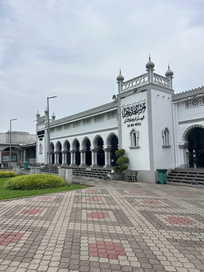 This is a medium-shot, eye-level, outdoor, daytime photograph of the Zahir Mosque in Alor Setar, Malaysia. The mosque is a large, white, Islamic religious building with a prominent tower. The architecture features arched walkways supported by columns, decorative latticework, and small domed finials on the tower. Arabic calligraphy and a date, "11-03-1912", are visible on the facade. In front of the mosque is a paved walkway with a distinctive pattern of interlocking grey and red tiles. To the left, there is a well-maintained lawn with manicured shrubs. A wooden bench and a potted plant are situated near the entrance. A green trash bin is visible to the right of the entrance. The sky is overcast and light grey, indicating a cloudy day. There are no discernible people in the immediate foreground, though shadows and faint figures suggest presence within the arched walkway of the mosque. The scene appears to be a street-level view of the mosque and its immediate surroundings.